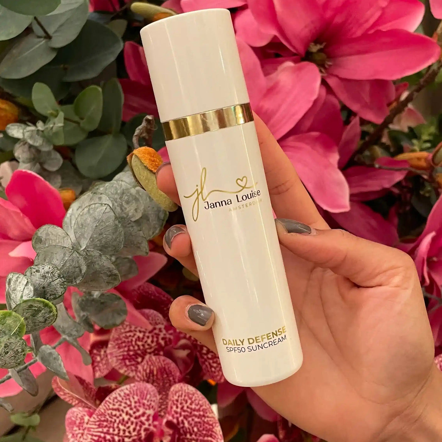 Hand met flesje SPF50 gezichtscrรจme van Janna Louise voor luxe huidverzorging met bloemen op de achtergrond