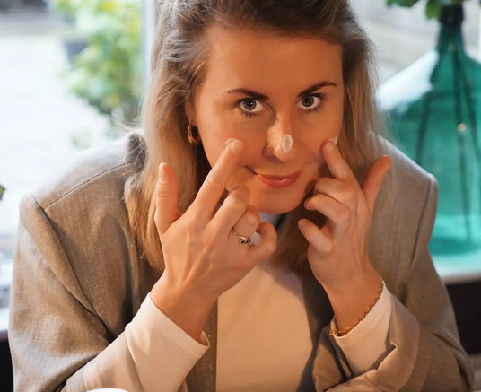 Vrouw brengt met vingers crèmepunten aan op haar gezicht als onderdeel van huidverzorgingsroutine in huiselijke setting.