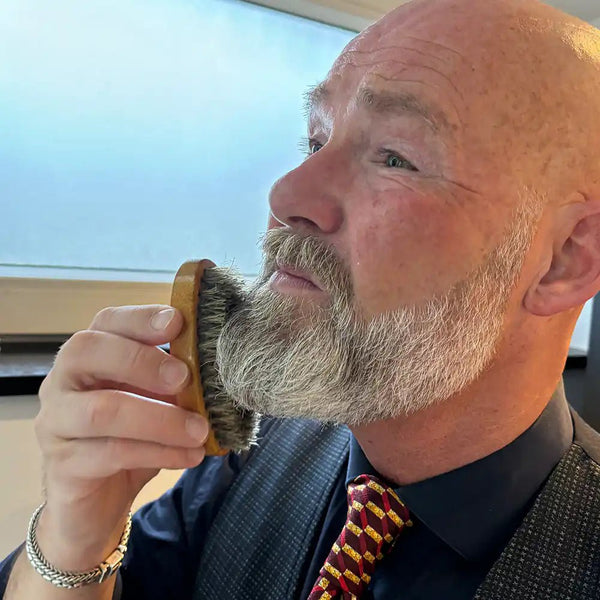 Man met grijze baard borstelt zijn gezicht met een houten baardborstel in een lichte, moderne omgeving.