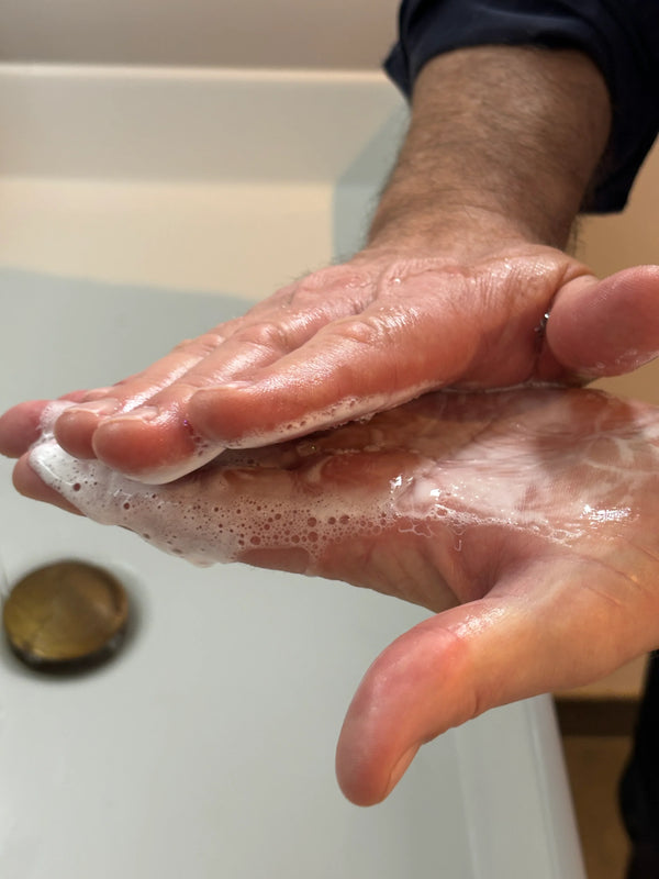 Close-up of hands with soap lather being rubbed together over a sink drain in a bathroom setting.