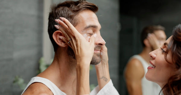 Vrouw brengt zachte gezichtsverzorging aan bij man in moderne badkameromgeving met spiegel op de achtergrond.