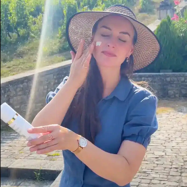 Vrouw met zonnehoed brengt dagcrème aan op gezicht in zonnige buitenomgeving met groene achtergrond.