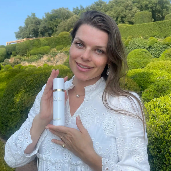 Vrouw met witte blouse toont luxe gezichtsverzorging in een groene tuinomgeving bij natuurlijk daglicht.
