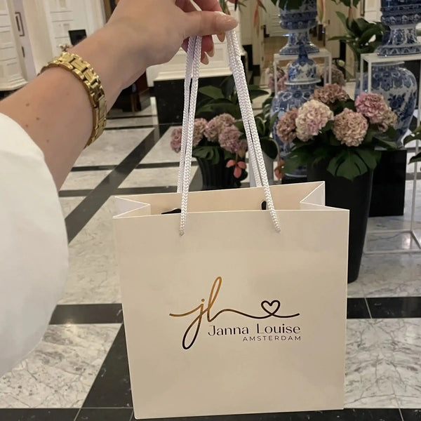 Hand met gouden horloge die een wit papieren tasje vasthoudt met logo van Janna Louise Amsterdam in elegante winkelomgeving.