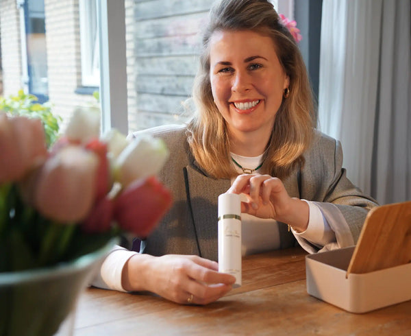 Vrouw in blazer toont een flesje huidverzorging aan een houten tafel met vaas bloemen op de voorgrond.
